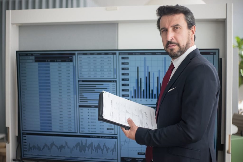 Businessman in a suit holds documents analyzing graphs on a screen, reflecting data analysis.