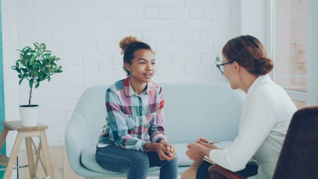 Cheerful African American girl is talking to psychologist sitting on sofa in modern loft style studio and smiling while specialist is listening to her and making notes.